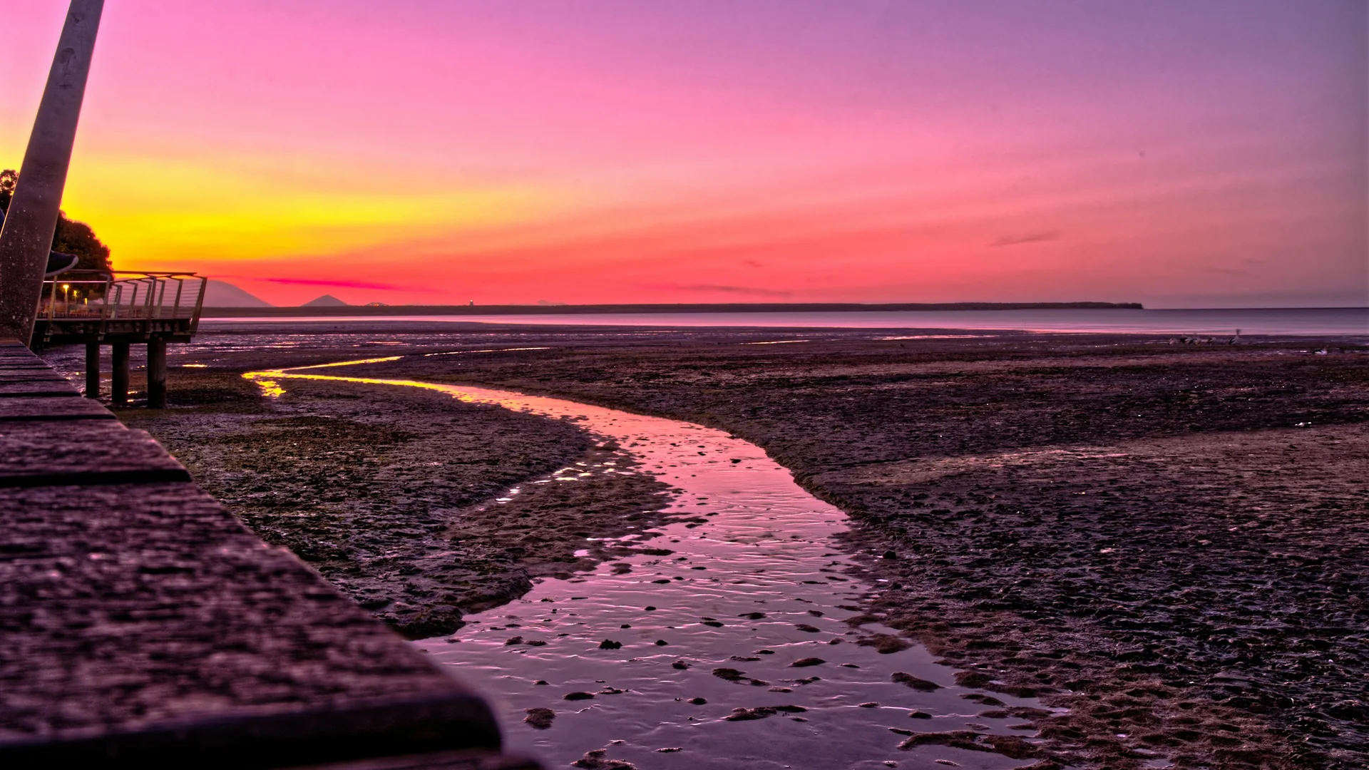 Un atardecer en tonos morados, rosas y dorados sobre el paseo marítimo de Cairns, con un canal de aguas tranquilas serpenteando entre las marismas junto a una pasarela de madera.