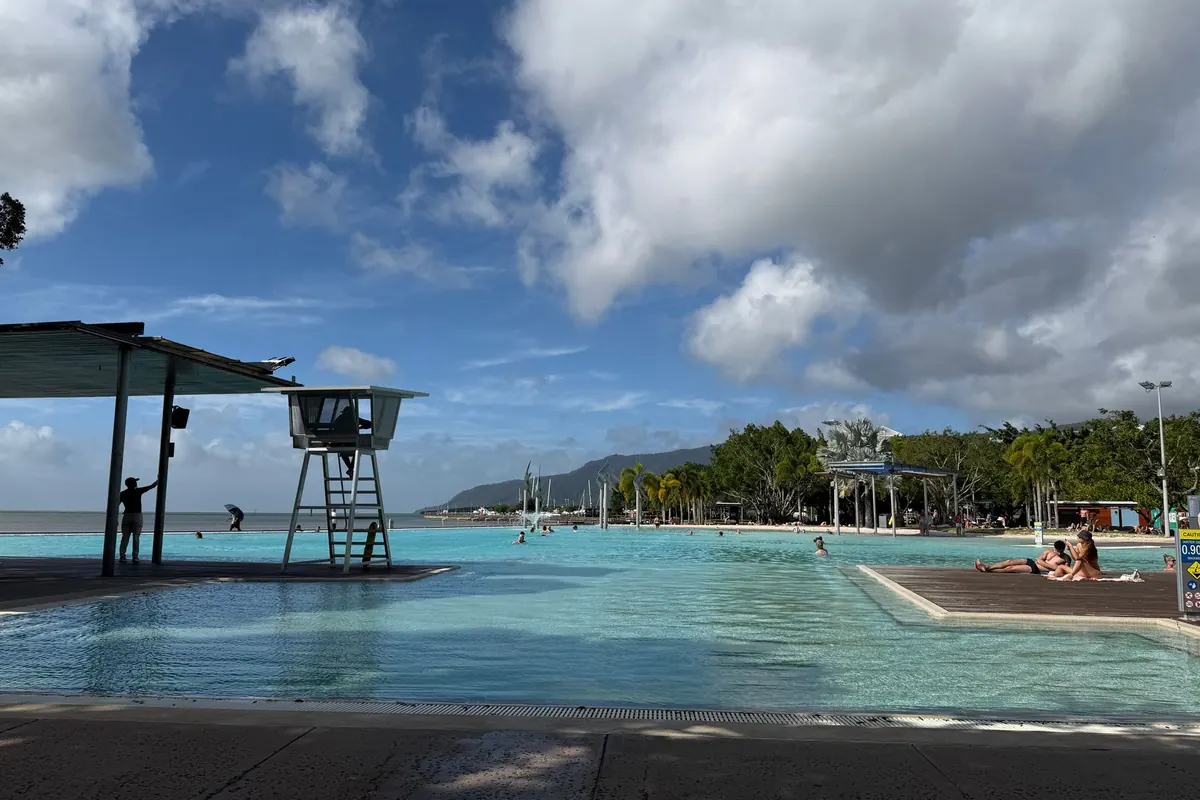 La laguna del Esplanade en Cairns, con una torre de socorrista junto a la piscina, bañistas en una terraza solar y colinas verdes a lo lejos.