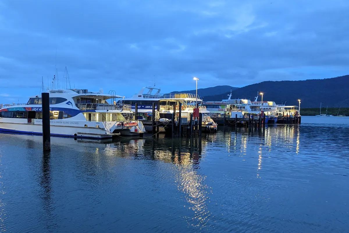 Barcos turísticos blancos y azules amarrados a un pontón en Cairns al atardecer, con sus luces reflejadas en aguas tranquilas y colinas al fondo