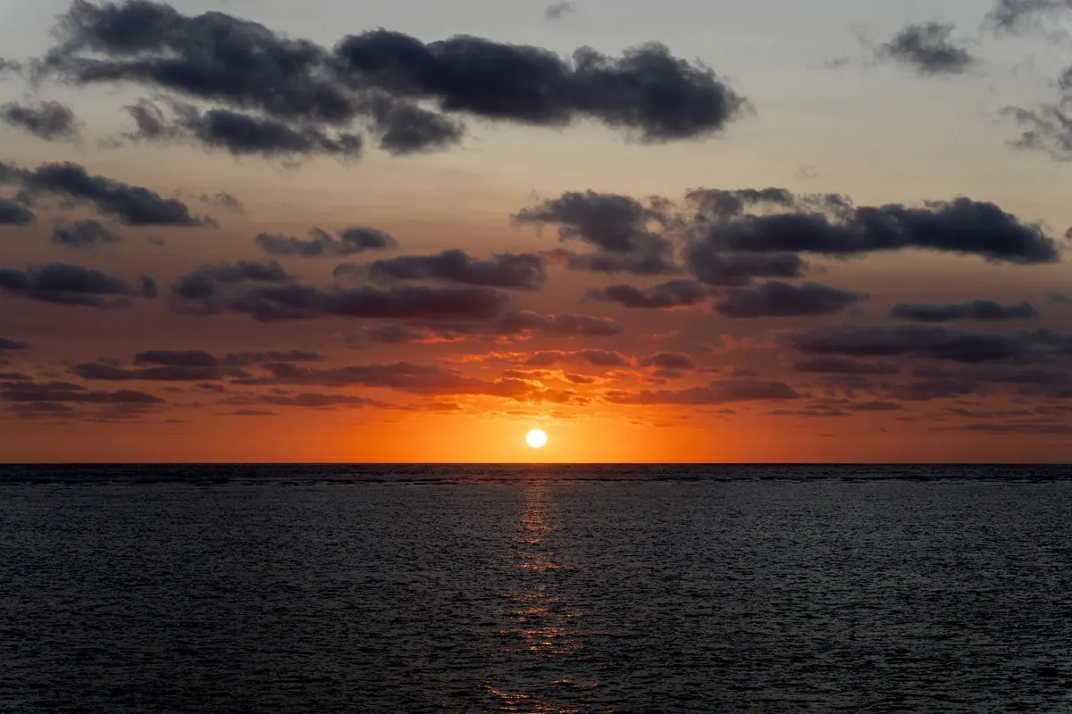 El sol hundiéndose en un mar oscuro frente a Cairns bajo franjas de nubes, con un resplandor anaranjado a lo largo del horizonte.
