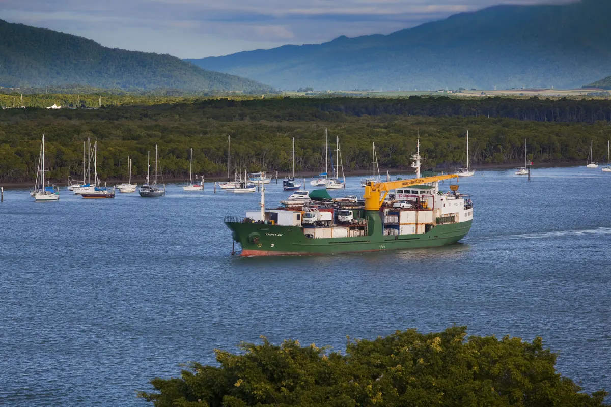 Un buque de carga verde con una grúa amarilla que pasa junto a veleros amarrados en Trinity Inlet, con una oscura línea de manglares y colinas boscosas al fondo